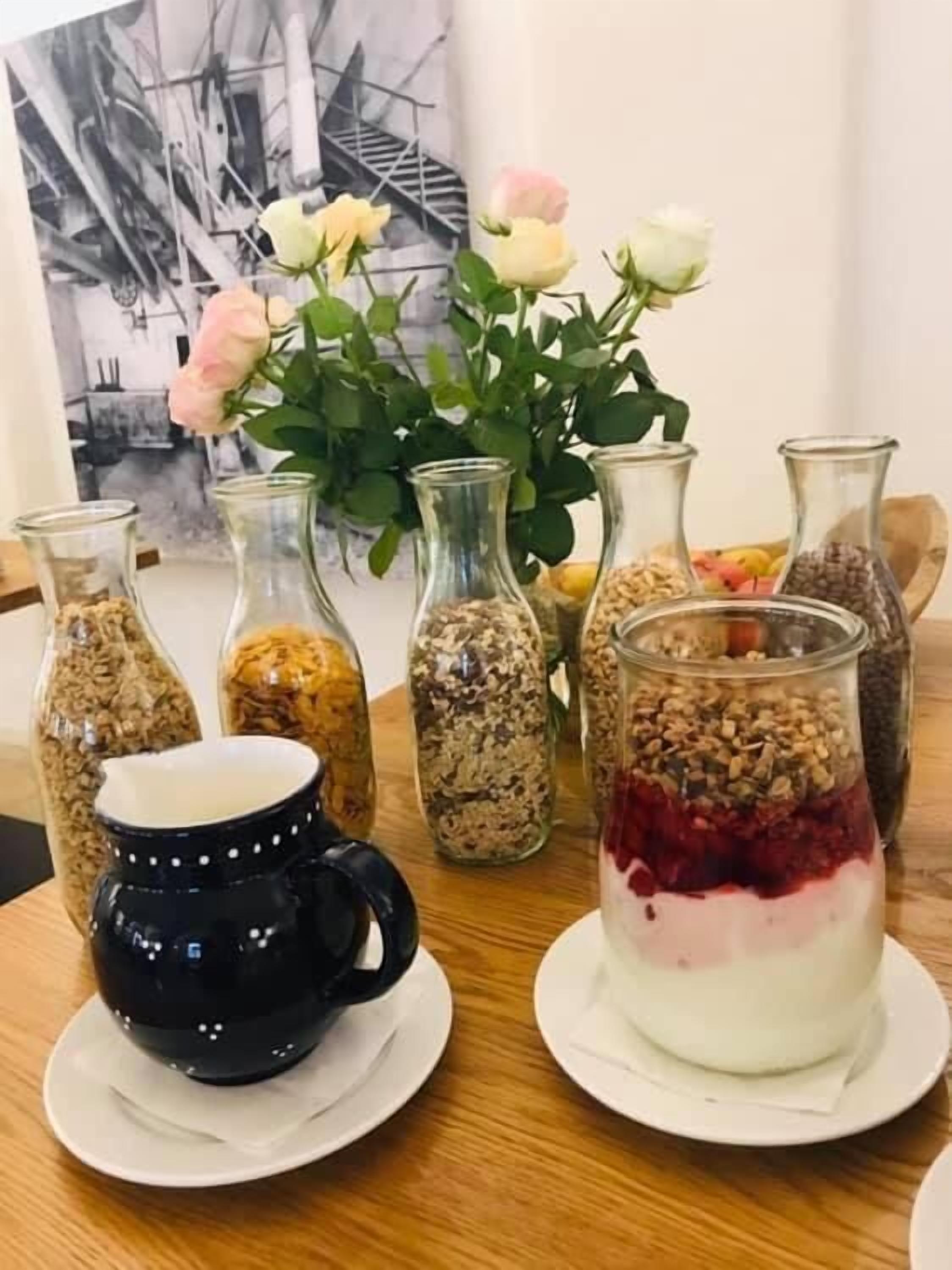 Una mesa con frascos de granola, una taza, un jarrón con flores y un plato con un postre en capas.