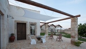 Een terras buiten met stenen vloer, witte stoelen, een tafel en een pergola.