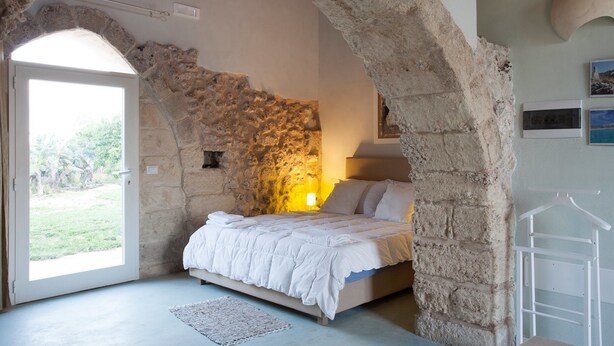 Slaapkamer met een stenen muur, een bed met wit beddengoed, een hanglamp en uitzicht op groen buiten.