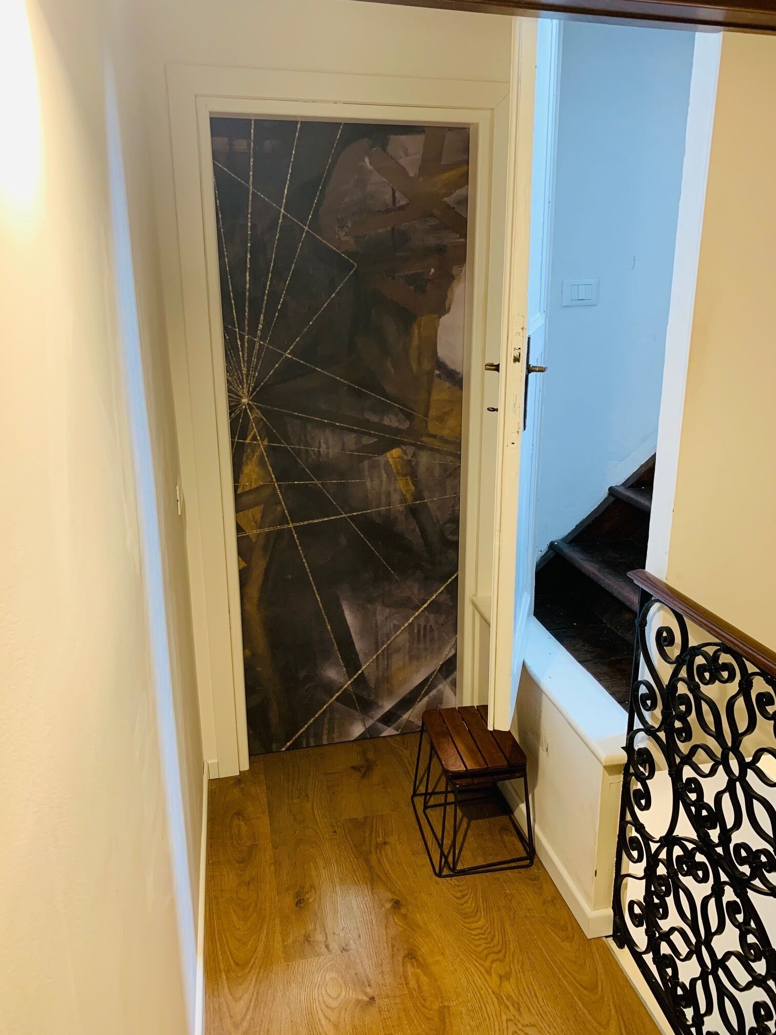 Una scala in legno con ringhiera in metallo nero che conduce a una stanza con una porta bianca decorata con un motivo geometrico.