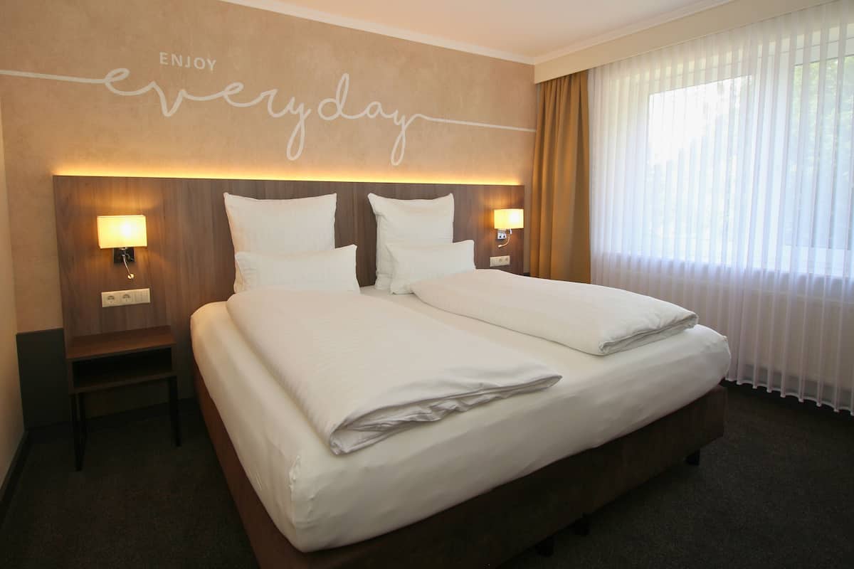 A hotel room with a large bed, two bedside tables with lamps, a headboard with a motivational quote, and a large window with sheer curtains.