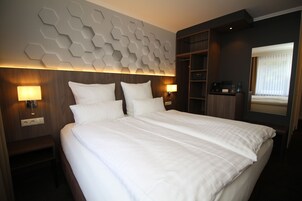Quarto de hotel com uma cama grande, duas luminárias de cabeceira, uma cabeceira de madeira e um design geométrico na parede.