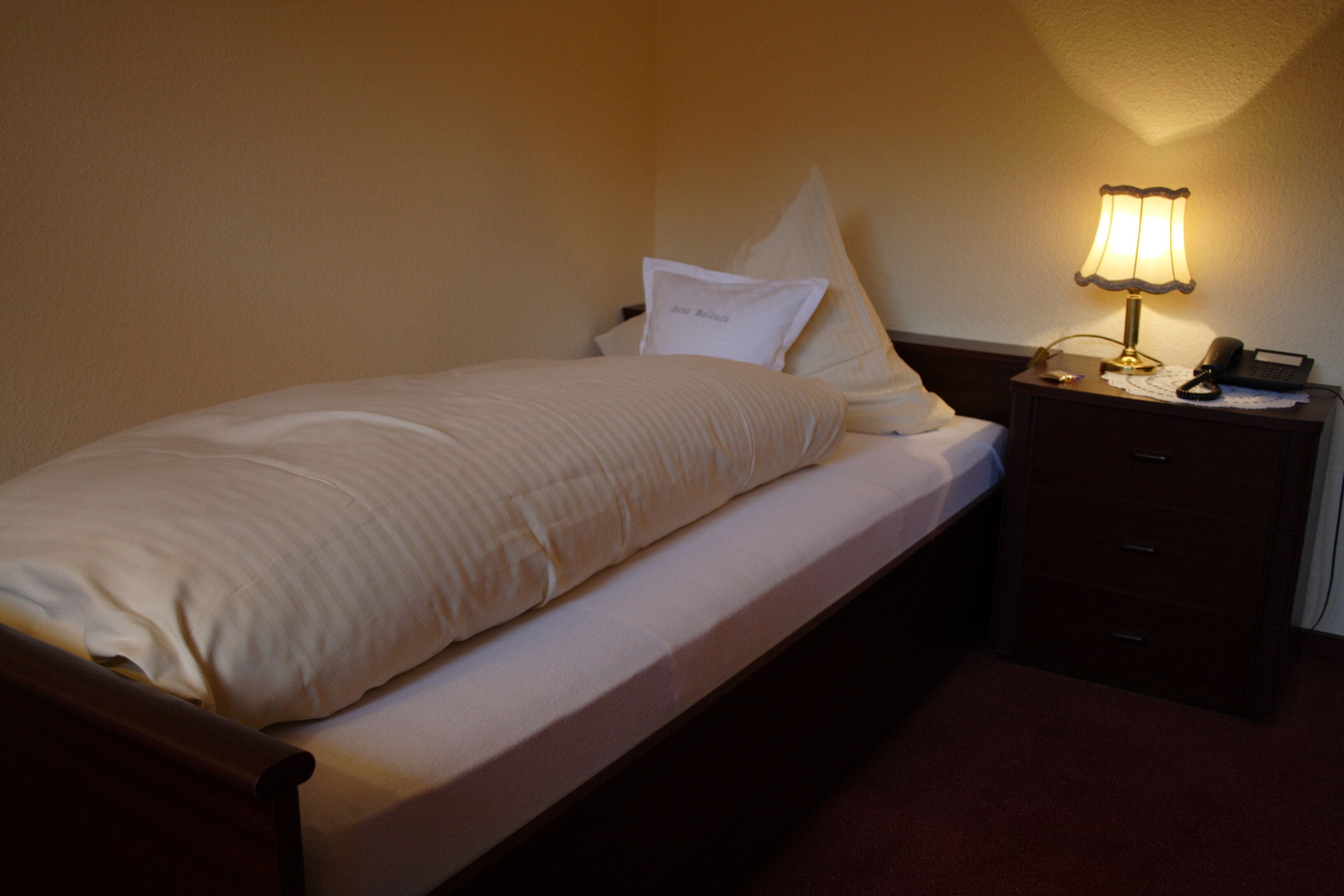 Una cama individual con edredón blanco y almohadas, una mesita de noche con lámpara y teléfono.