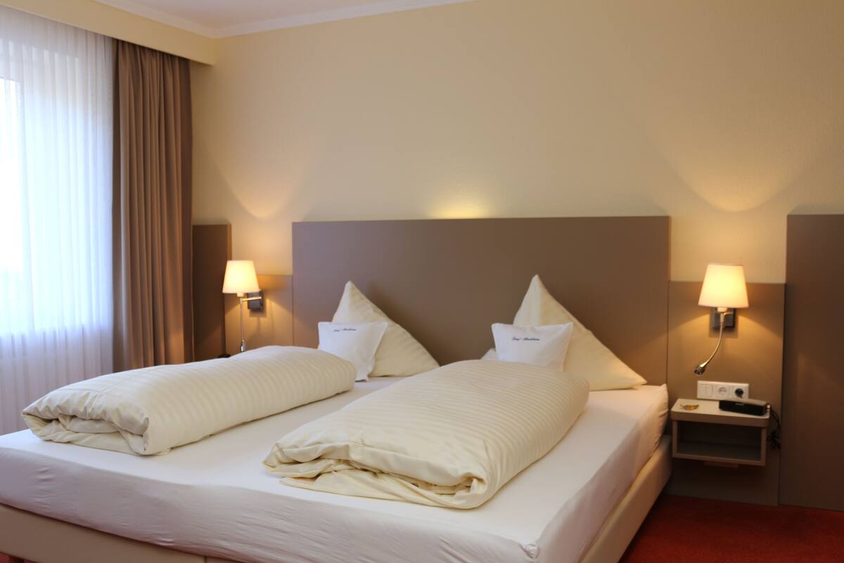 A hotel room with two beds, each with white linens and pillows, a nightstand with a lamp, and a window with curtains.