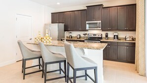 A modern kitchen with dark wood cabinets, a granite countertop, a stainless steel microwave, a refrigerator, and a coffee maker. There are three bar stools with grey upholstery. - Delightful Home with Private Pool Oasis in Encore (Kissimmee)
