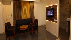 A modern living room with a sofa, two armchairs, a coffee table, a flat-screen TV mounted on a wooden wall, and a door with curtains. - DFRONT Golden Palace (Namakkal)