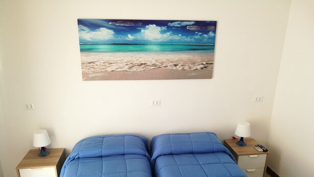 A room with two beds, a painting of a beach scene, and a nightstand with a lamp and a remote control. - B&B Laura e Chiara (Trapani)