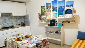 A small, cozy kitchen with a dining table set for two, featuring a colorful tablecloth and a variety of dishes. A coffee machine and a shelf with kitchen essentials are visible. - B&B Laura e Chiara (Trapani)