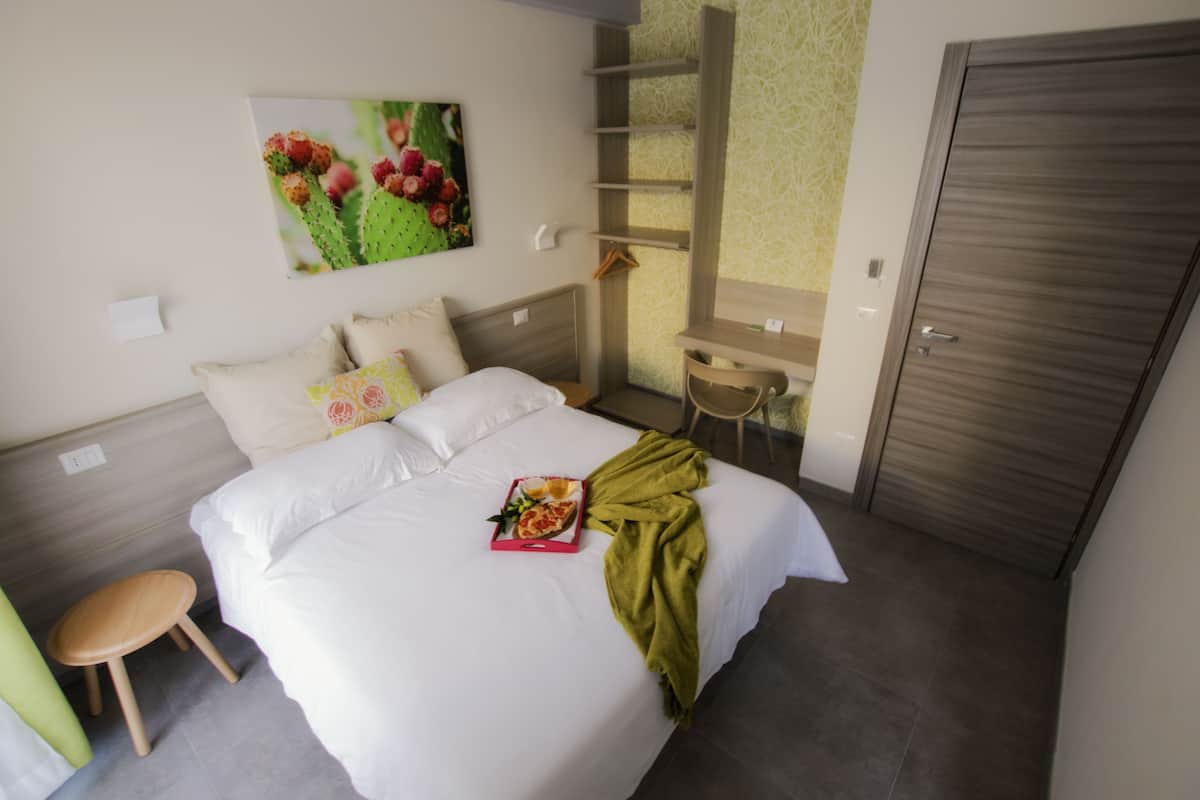A neatly made bed with pillows, a tray with food, and a green blanket. There is a chair and a shelf with a hanging item.