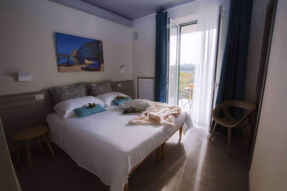 A hotel room with a large bed, a chair, a painting on the wall, and a view of the outdoors through the window.