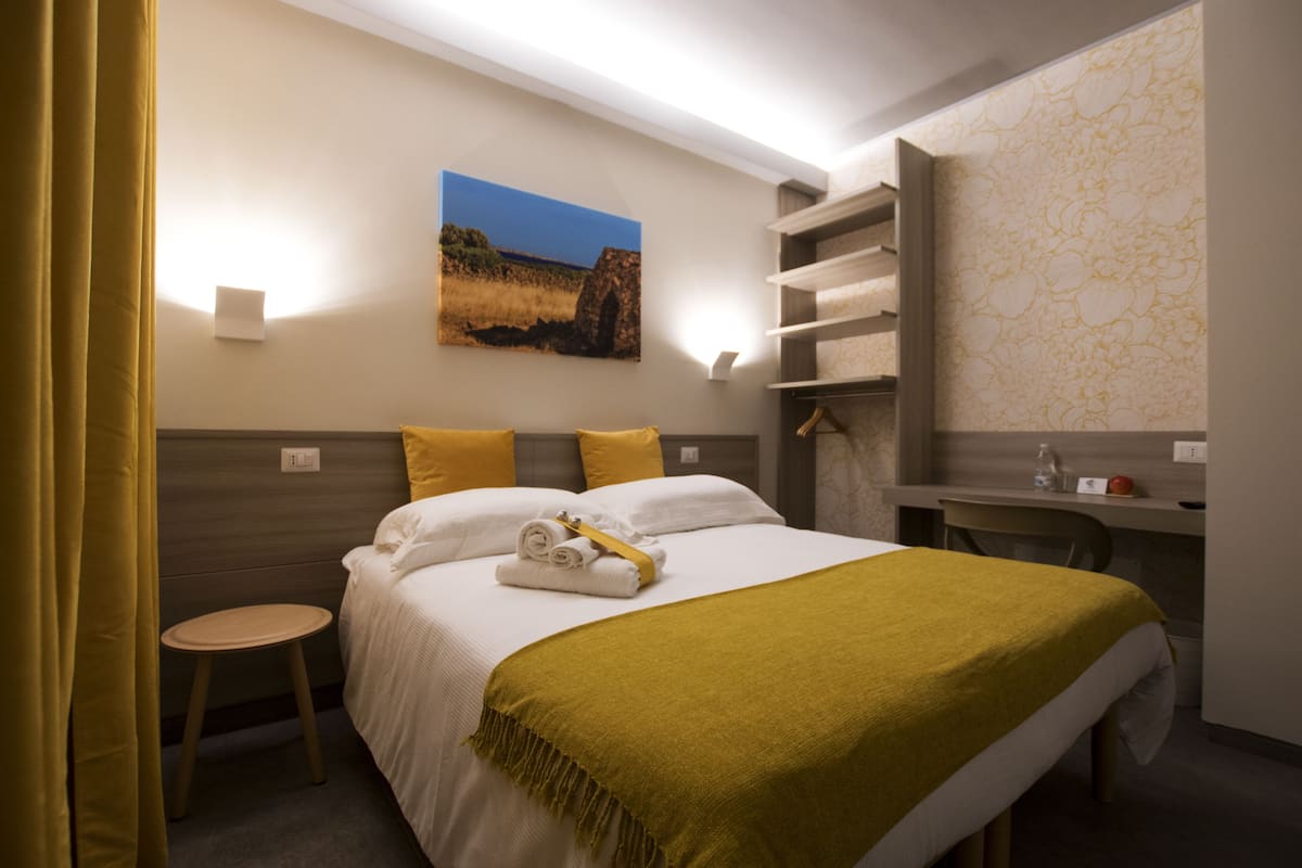 A hotel room with a bed, a desk, a chair, and a small stool.