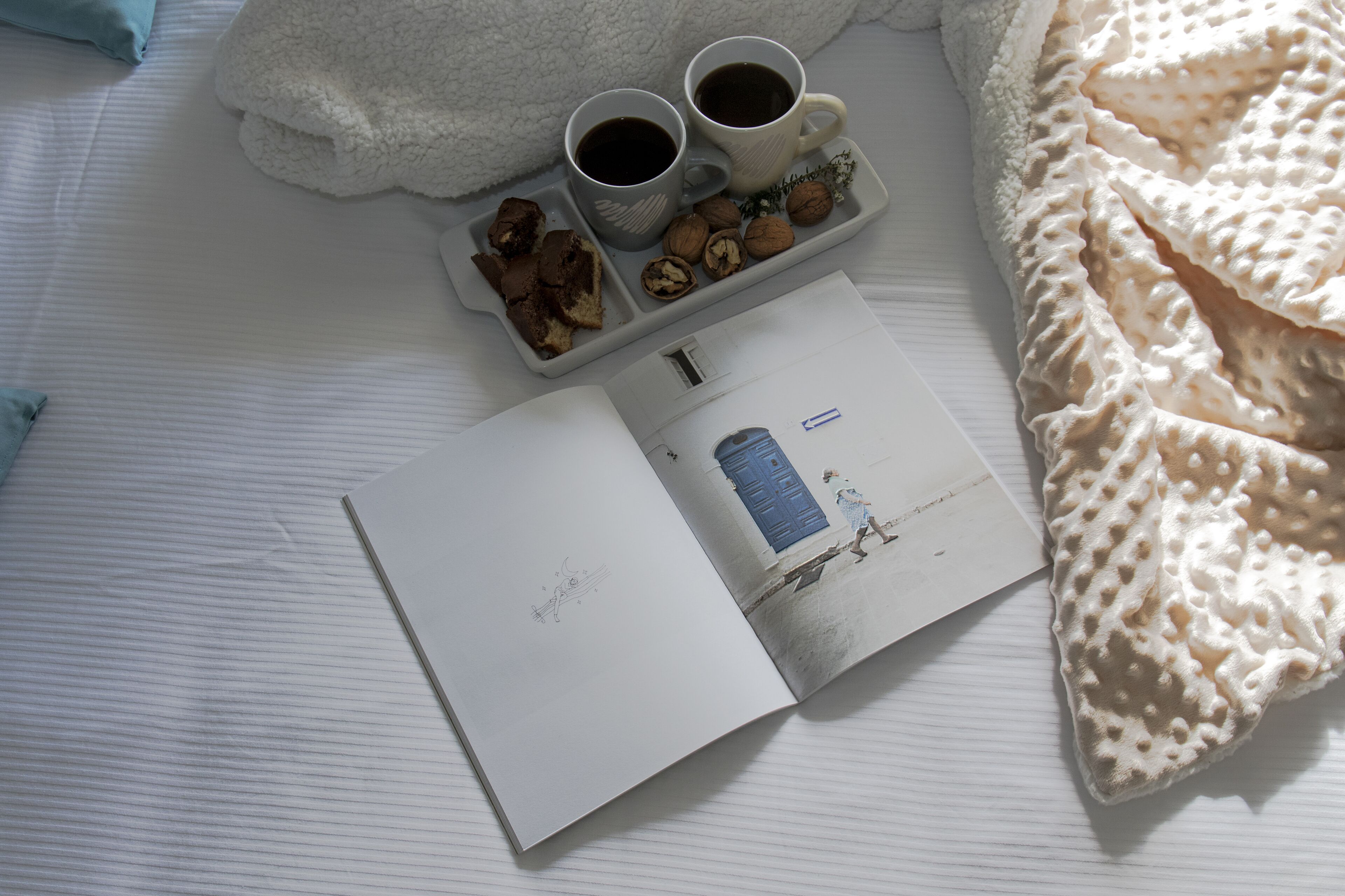 Una cama con una manta blanca, una bandeja con dos tazas de café, un libro y algunos bocadillos.