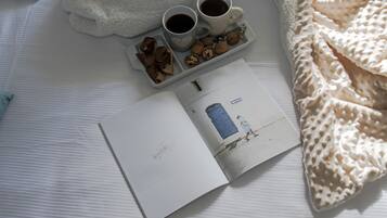 Una cama con una manta blanca, una bandeja con dos tazas de café, un libro y algunos bocadillos.