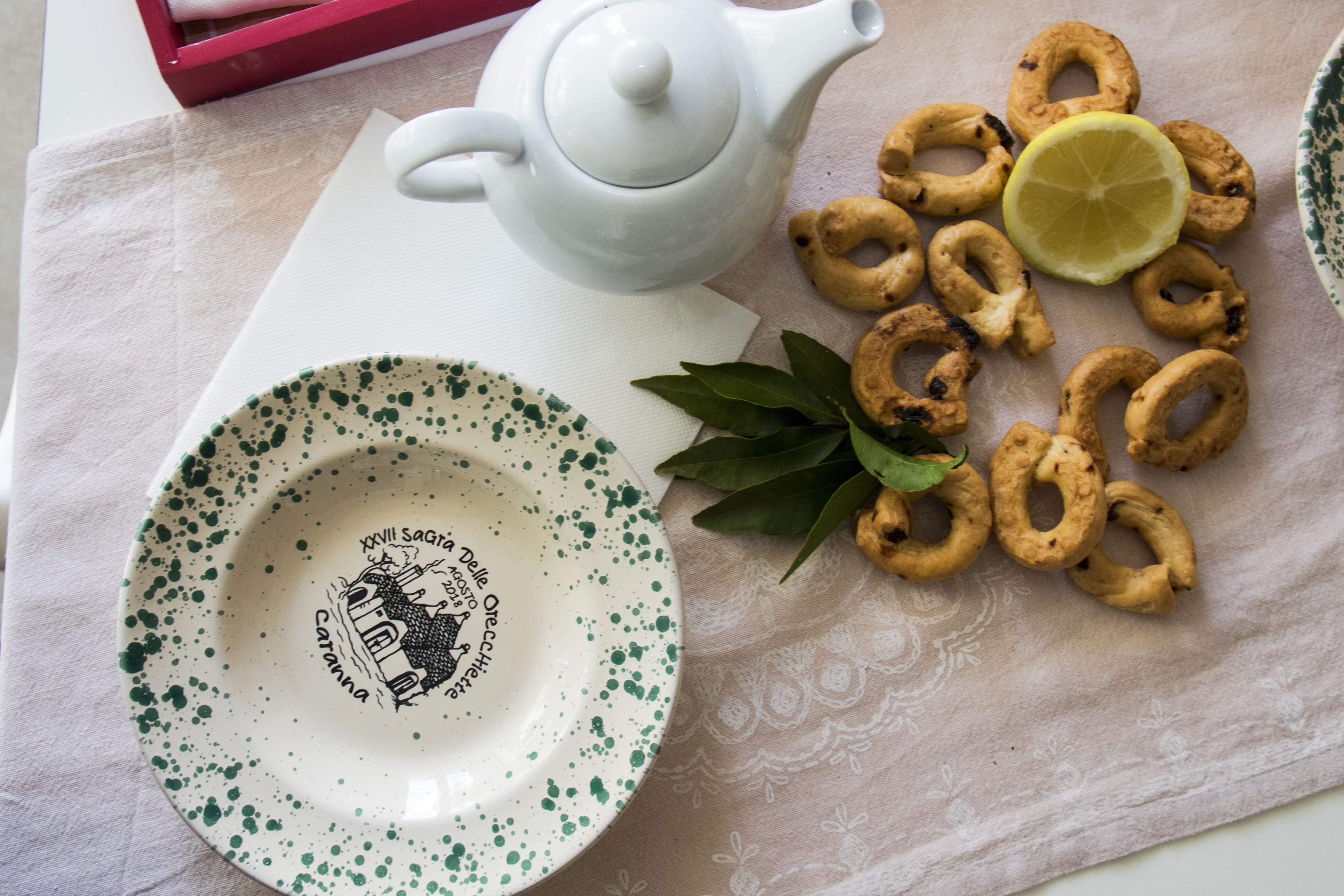 Una tetera blanca, un plato con motas verdes y galletas con forma de anillo sobre un mantel.