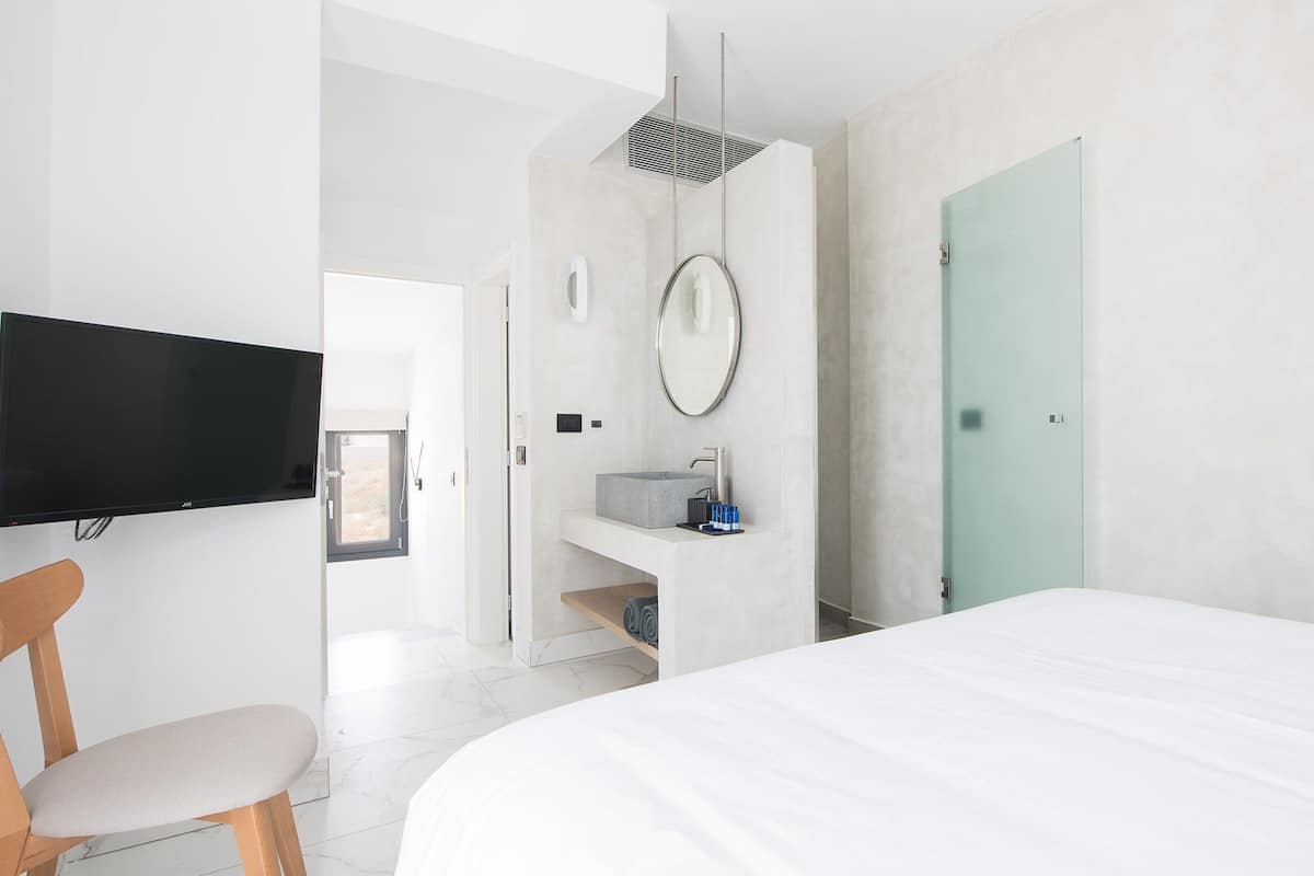 A modern bedroom with a large bed, a TV mounted on the wall, a bathroom with a sink and mirror, and a door leading to another room.