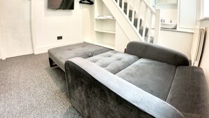 Une pièce avec un canapé gris, un téléviseur fixé au mur, un escalier et une petite étagère contenant des articles de toilette.