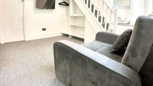 Un salon avec un canapé gris, un escalier blanc, un téléviseur fixé au mur et une petite étagère avec un miroir.
