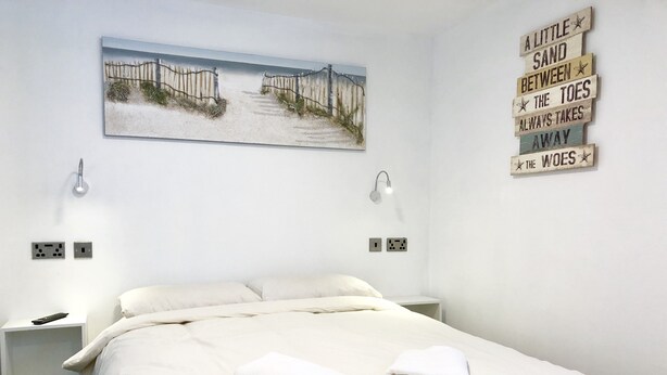 Un lit bien fait, avec du linge de lit blanc et deux serviettes pliées, un tableau représentant une scène de plage et un panneau mural affichant une citation inspirante.