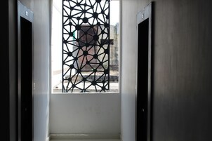 Vista del interior de una habitación de hotel con una ventana de diseño geométrico, una pared blanca y una puerta a la derecha.