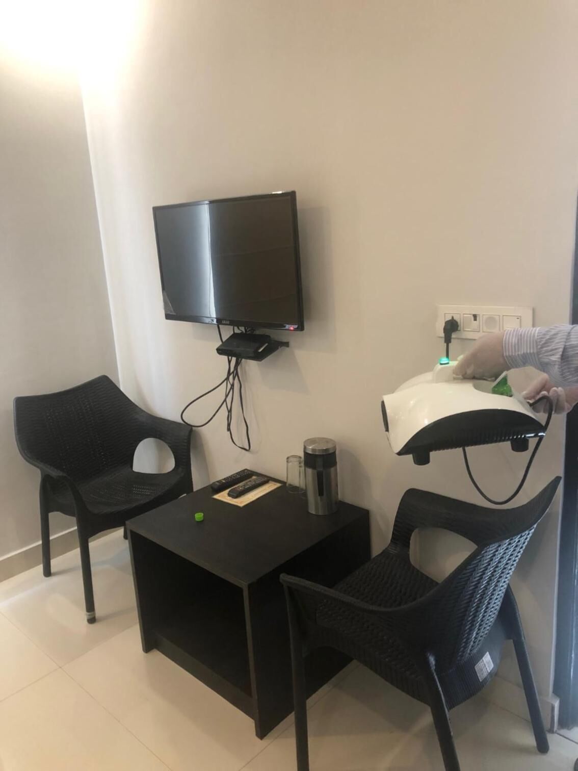 Quarto pequeno com televisão de tela plana presa na parede, uma mesa pequena com um controle remoto, um termo e duas cadeiras.