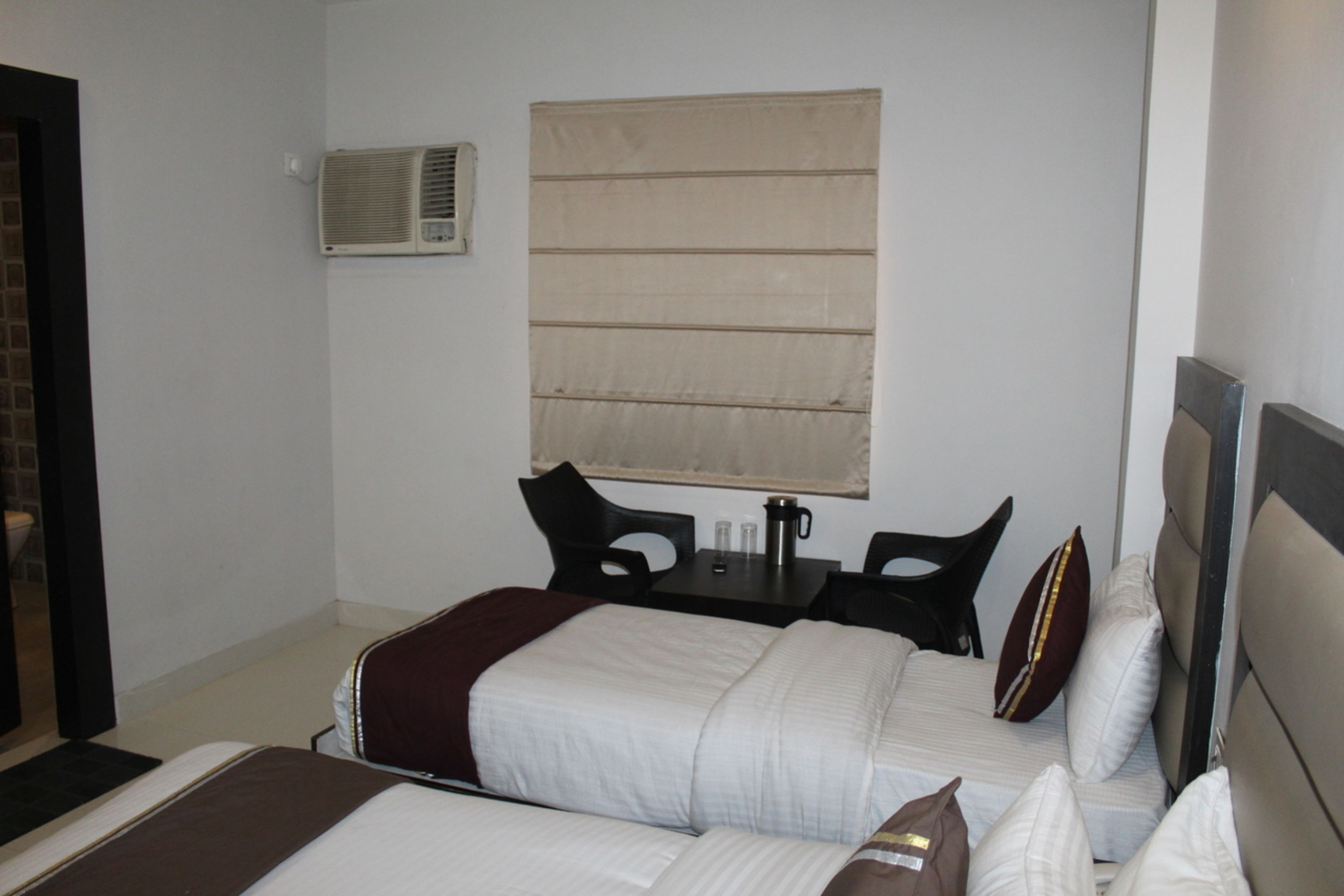 Habitación de hotel con dos camas, una mesita con sillas, un ventanal con persianas y un equipo de aire acondicionado en la pared.