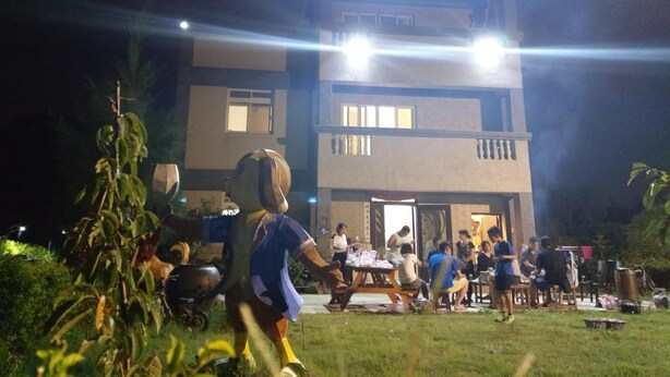 Un edificio de varios pisos y techo plano, rodeado de un espacio verde con gente reunida alrededor de mesas.
