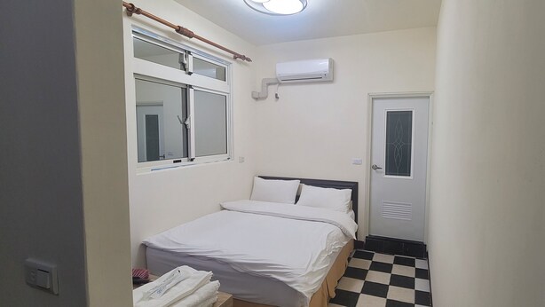 A small, clean room with a single bed, a window with a curtain, a door with a window, and an air conditioning unit on the wall. - Kinmen Good Places (Jinning)