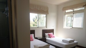 Habitación de hotel con dos camas, cada una con una almohada y una toalla doblada. Ventanas con cortinas y suelo de baldosas.
