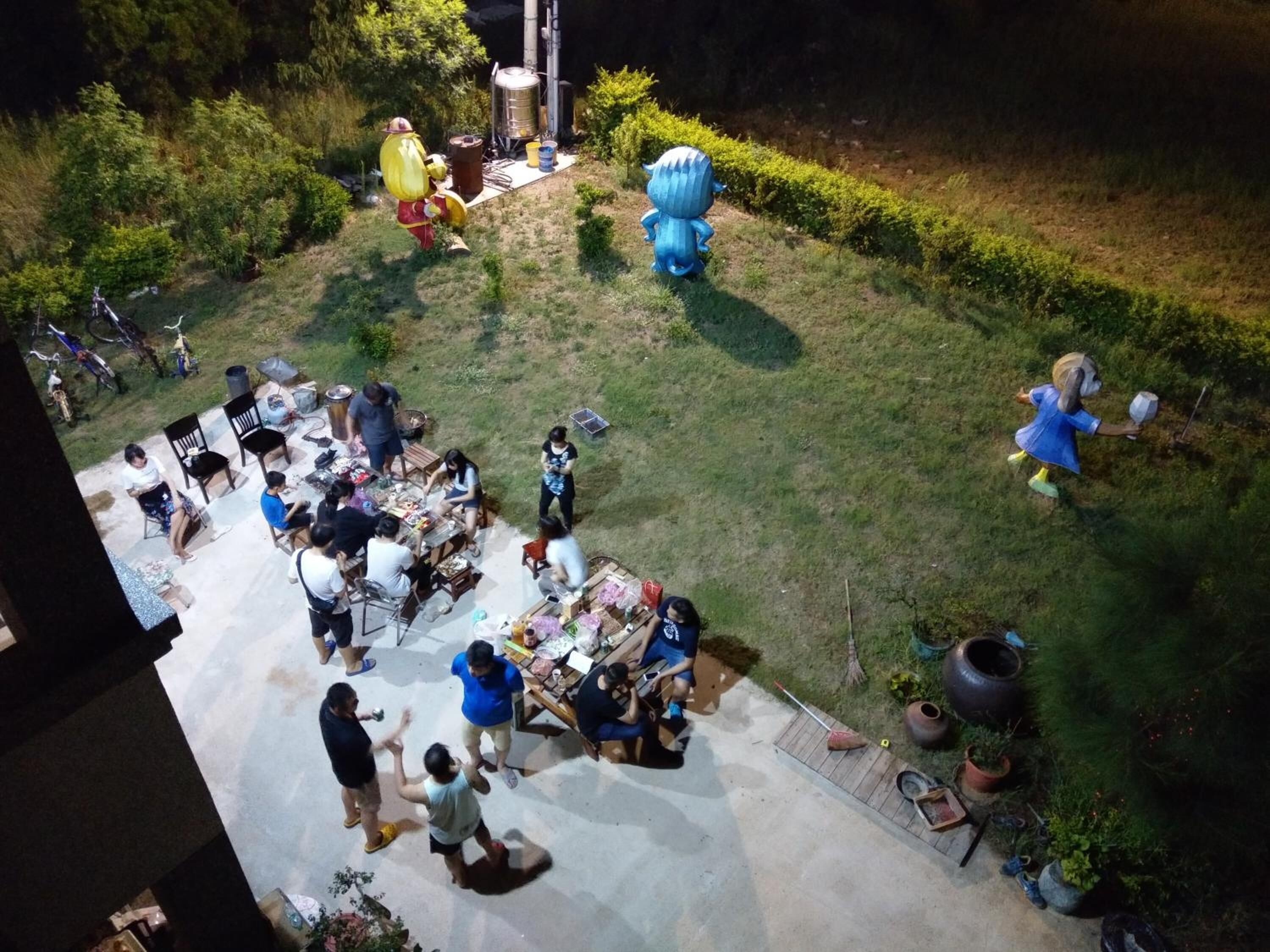 A courtyard with a group of people gathered around a table, possibly dining or socializing. There are various items and decorations scattered around, including a large blue sculpture and a yellow figure.