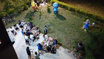 A courtyard with a group of people gathered around a table, possibly dining or socializing. There are various items and decorations scattered around, including a large blue sculpture and a yellow figure.