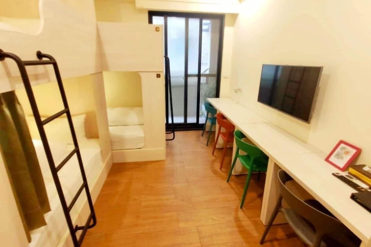 A compact room with a bunk bed, a desk with chairs, a television, and a sliding door leading to a balcony.