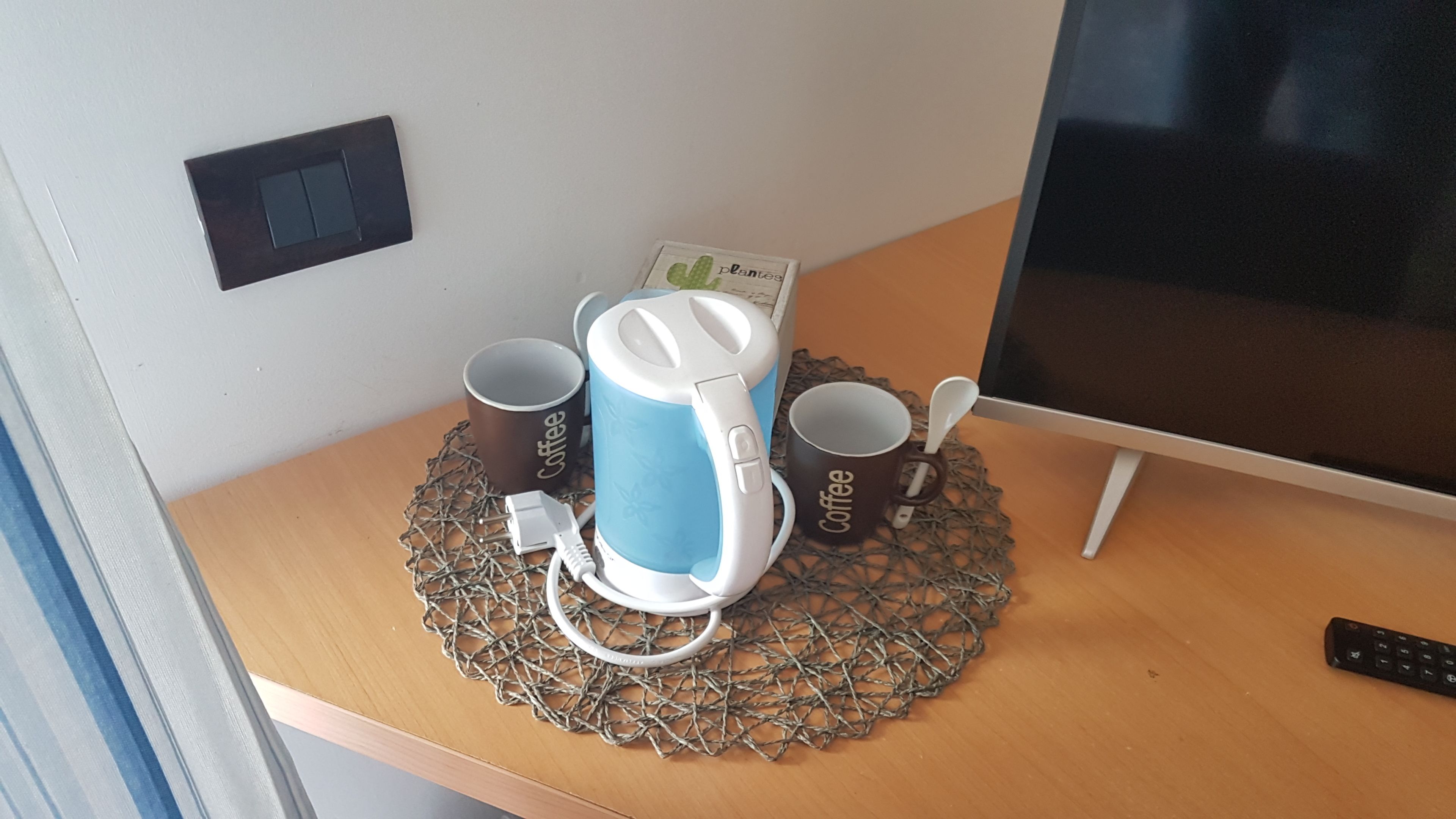A desk with a coffee maker, two coffee mugs, a remote control, and a computer monitor.