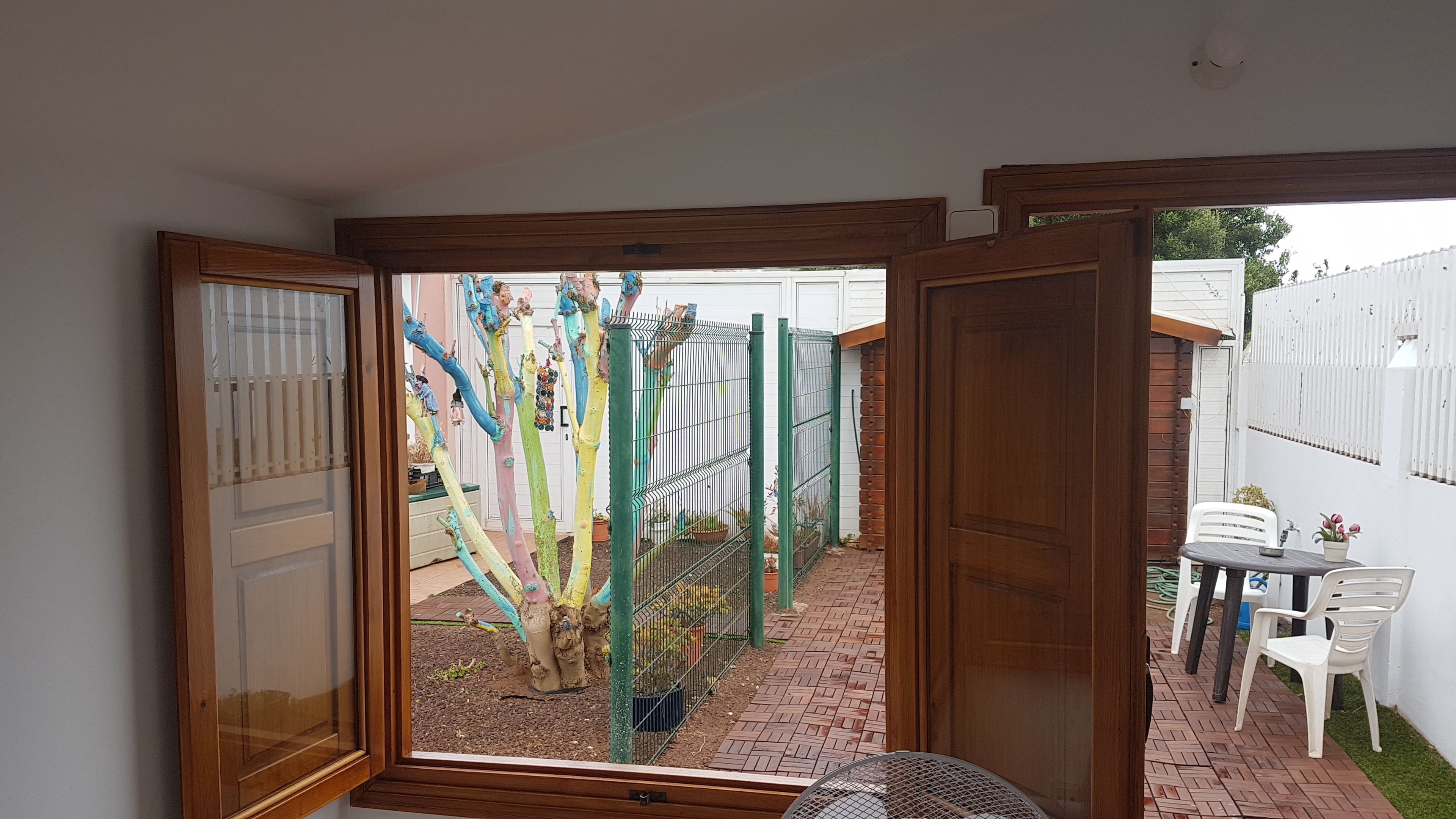 Uitzicht door een deuropening op een achtertuin met een geplaveid pad, een houten deur, een wit hekwerk en een tafel met stoelen.