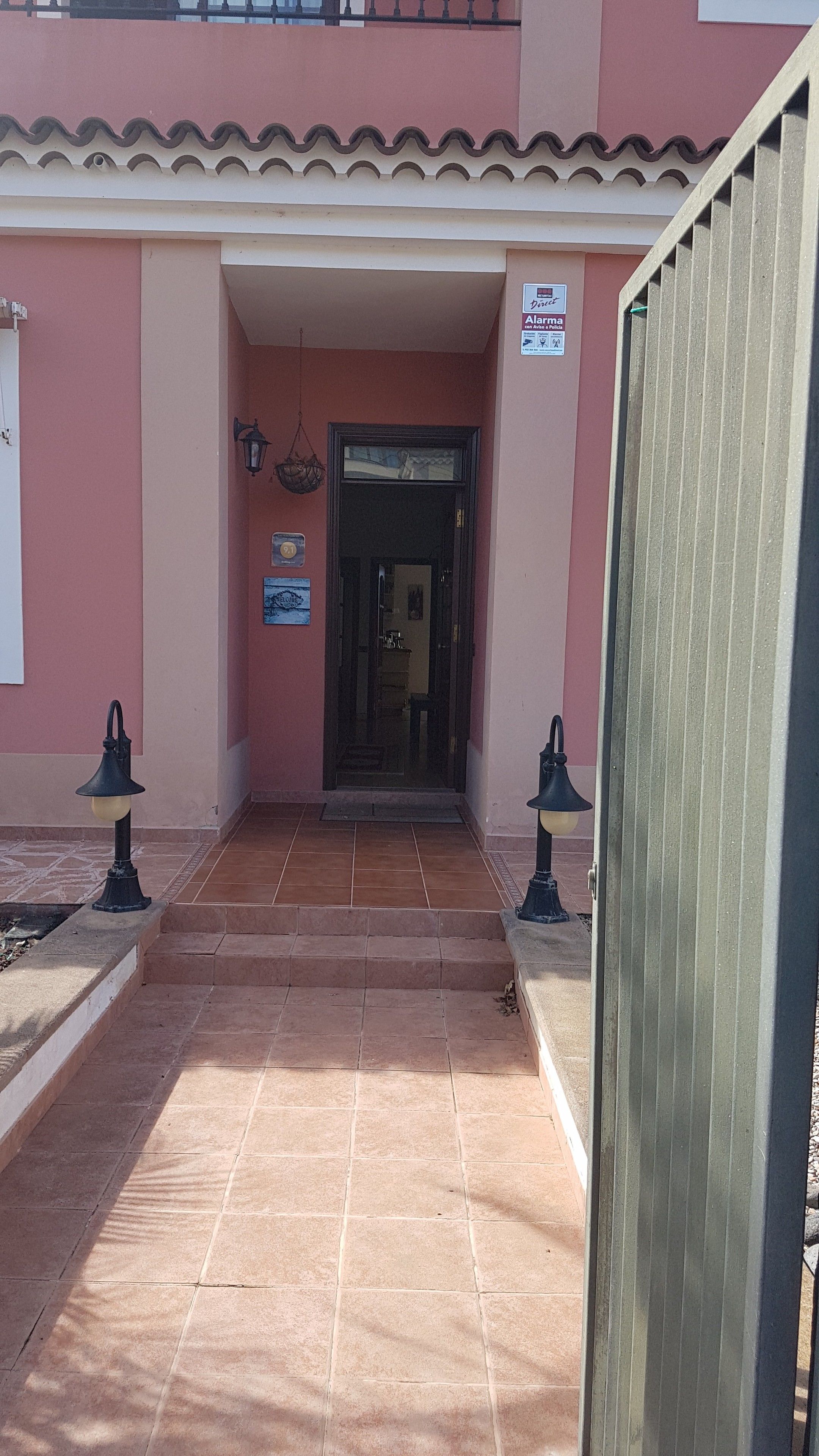 Una entrada de edificio con un camino de baldosas que conduce a una puerta, dos faroles en la pared y un letrero.