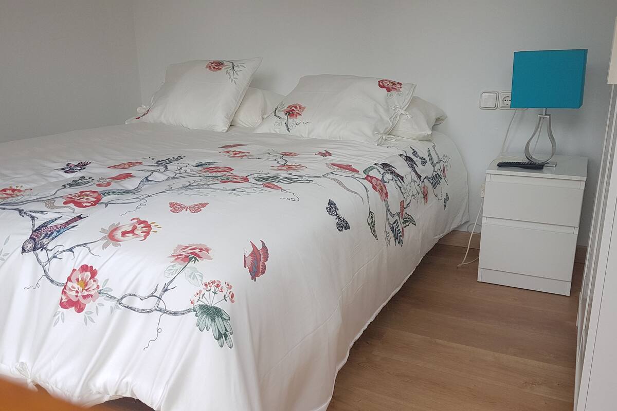 A neatly made bed with floral bedding, a bedside table with a lamp, and a wooden floor.