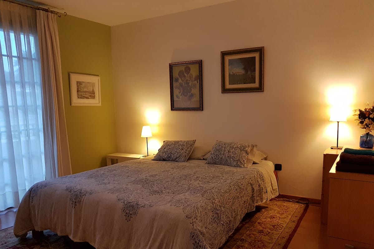 A bedroom with a large bed, two bedside lamps, a nightstand, a dresser, and a window with curtains.