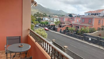 A balcony with a table and chairs, overlooking a street with houses and a mountain in the background.