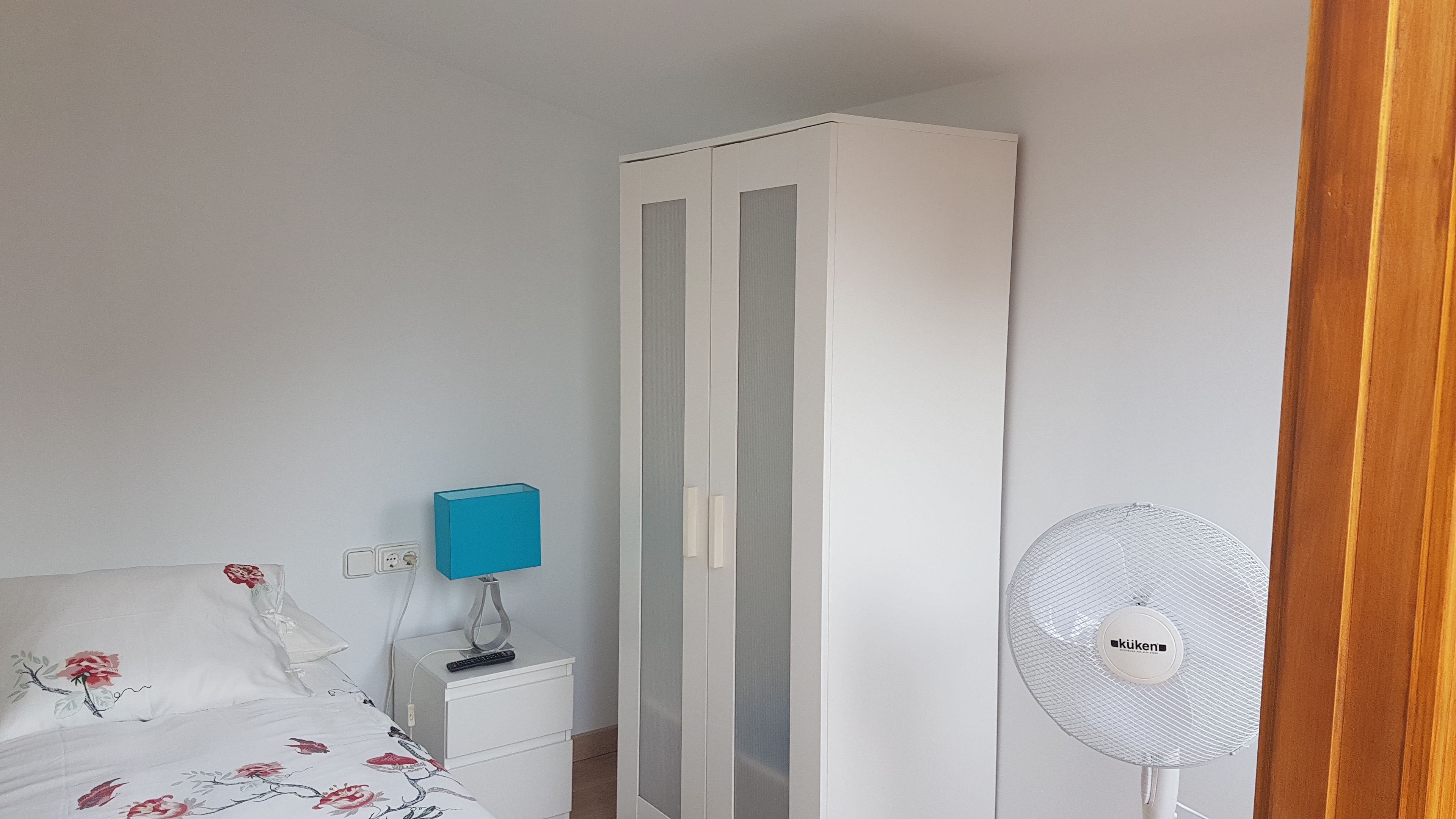 A bedroom with a bed, a nightstand, a wardrobe, and a standing fan.
