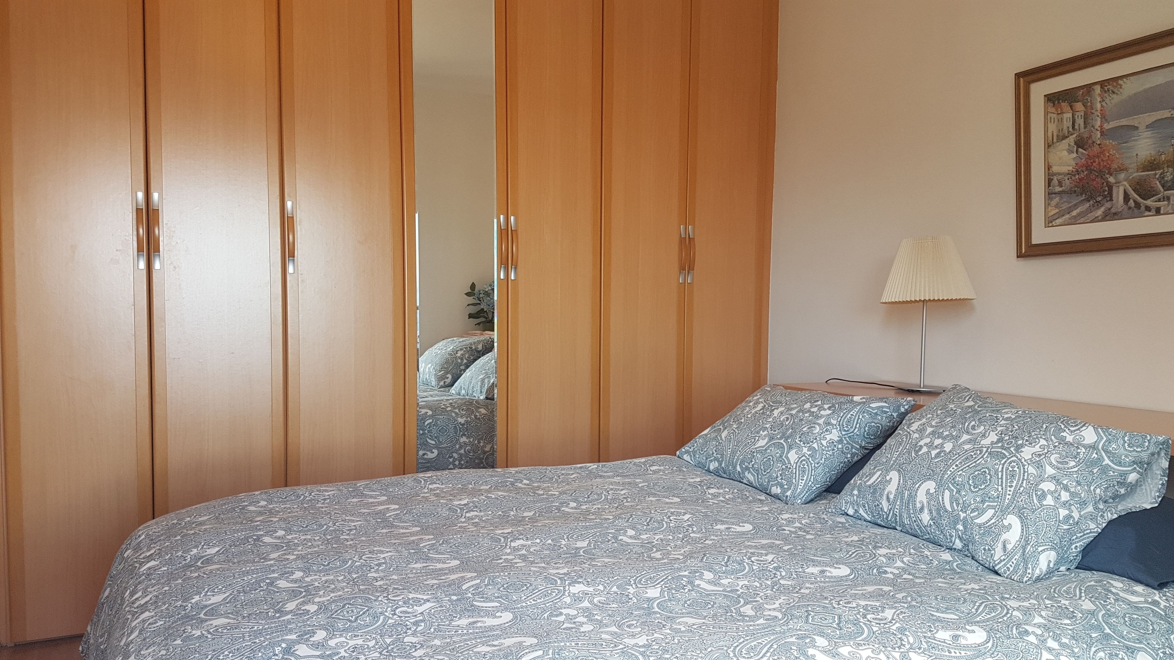 A bedroom with a large bed, patterned bedding, a wooden wardrobe, and a painting on the wall.