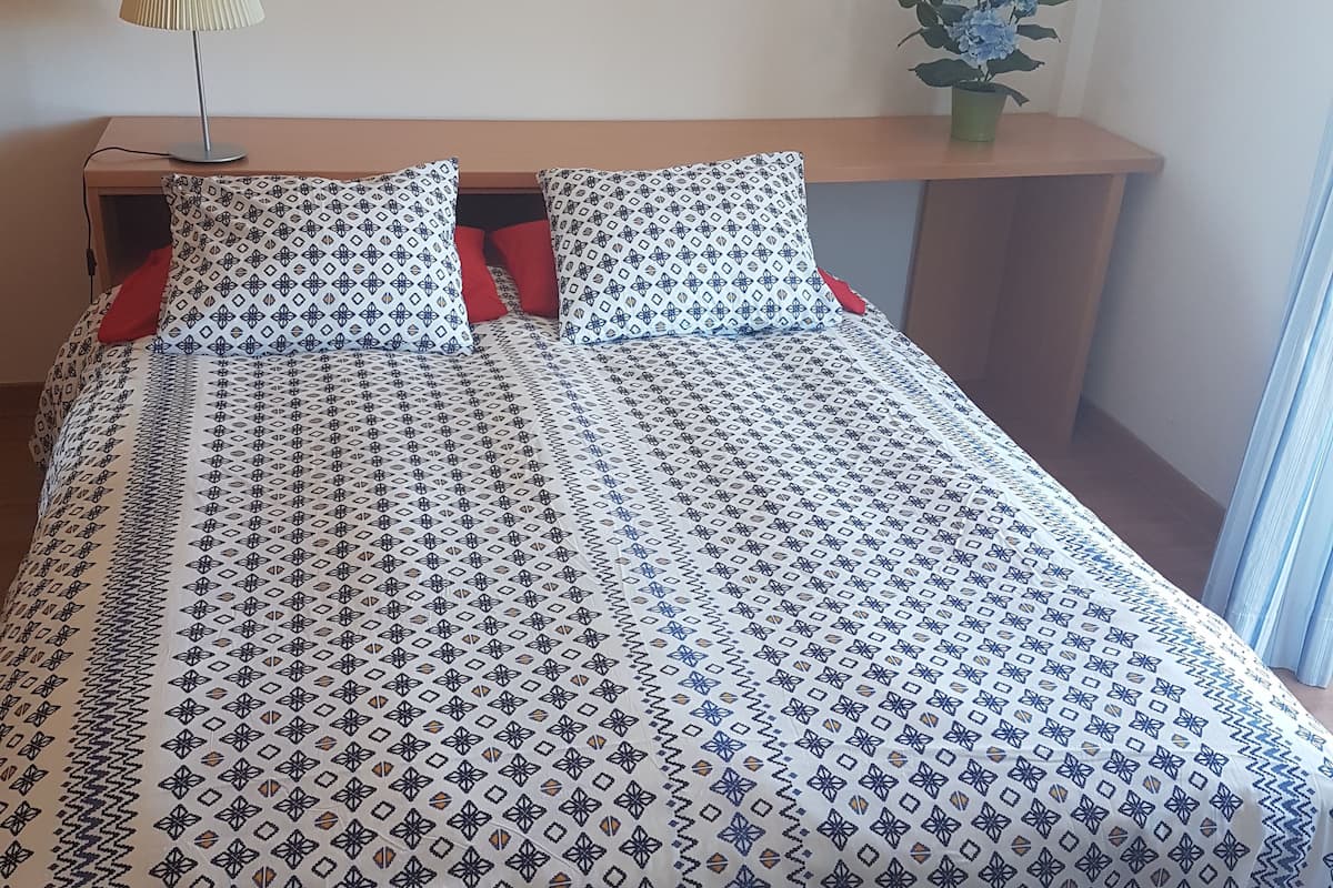 A neatly made bed with patterned bedding, a bedside lamp, a framed picture on the wall, and a small plant on a nightstand.