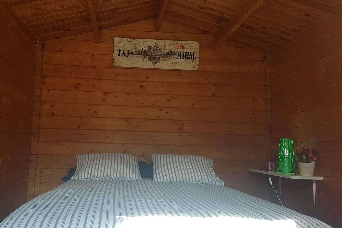 A wooden cabin room with a bed, striped pillows, a small shelf with a green cup, and a sign on the wall.
