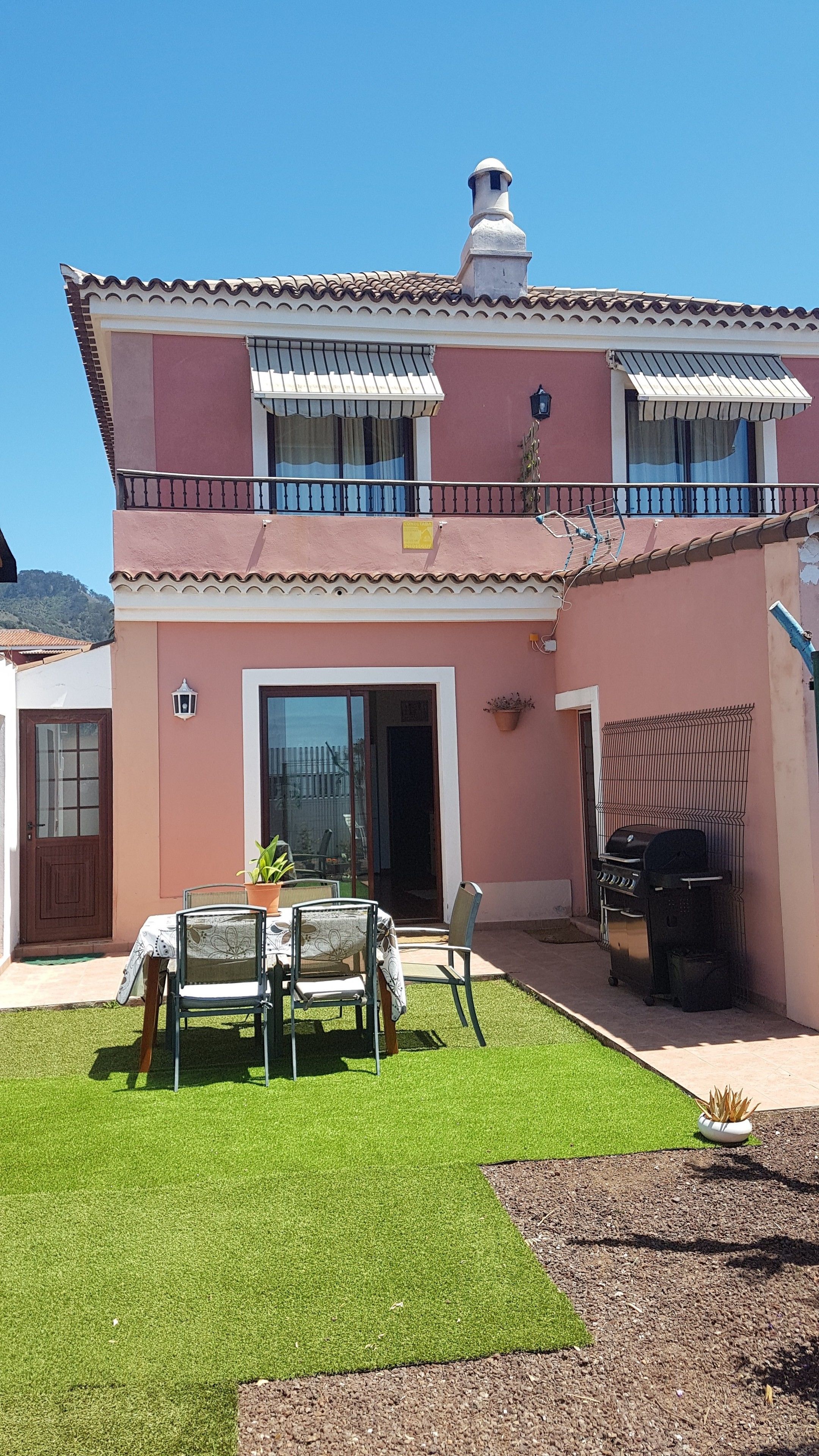 Een tweelaags huis met een balkon, een terras met tafel en stoelen, en een barbecue.