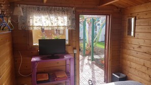 A wooden cabin room with a television on a purple stand, a window with a floral curtain, and a door leading outside. - Chalet Anagato (Tegueste)