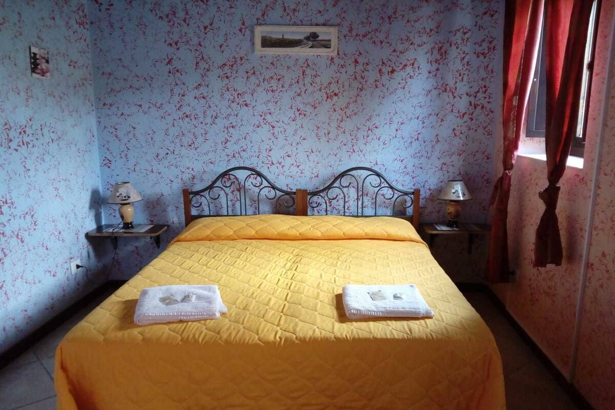 Um quarto com uma cama, duas mesas de cabeceira, uma lâmpada em cada uma, uma janela com cortinas vermelhas e uma parede com padrão floral.