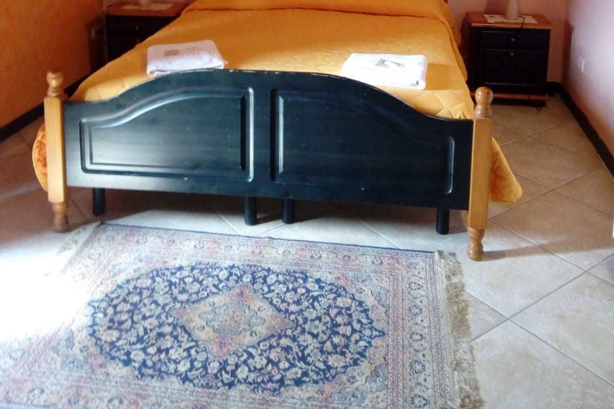 Uma cama com cabeceira escura, duas mesas de cabeceira com abajures, um tapete e uma pintura de pôr do sol na parede.