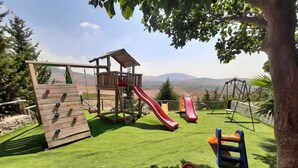 Un parque infantil con toboganes y estructuras para escalar sobre césped artificial, rodeado de árboles y un paisaje montañoso.