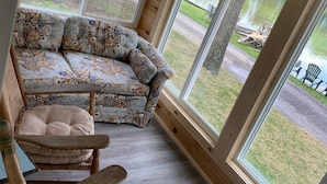 Ein gemütliches Zimmer mit Holzboden, einer blumigen Couch und einem Schaukelstuhl. Große Fenster bieten Blick auf eine Wiese und einen Steg.