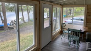 Eine Hütte mit großen Fenstern mit Blick auf einen See, einem Essbereich mit Stühlen und einem Tisch, und ein Geschwindigkeitsbegrenzungsschild ist draußen sichtbar.