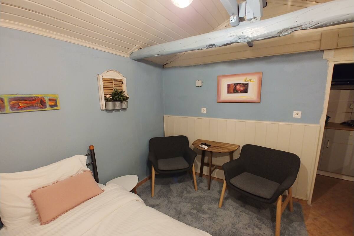 A small, cozy room with a bed, two chairs, a small table, and a window with shutters.