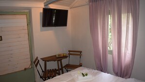 Una habitación con una cama, una silla de madera, una mesita con fruta, un televisor y una ventana con cortinas.